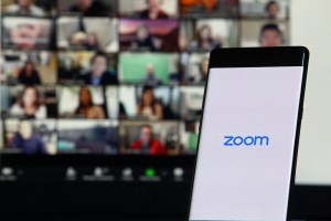 Foto de celular na tela inicial do aplicativo do Zoom, com computador conectado em teleconferência ao fundo