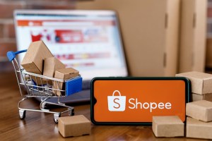 Foto de celular com logo da Shopee, carrinho de supermercado com pacotes em miniatura ao lado e um computador aberto no site da Shopee ao fundo