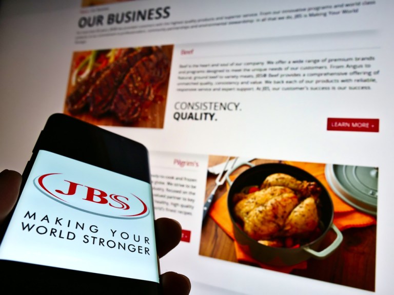 Celular com logo da JBS e site da empresa no fundo