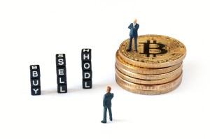 Arte com duas pessoas, sendo um em cima de uma pilha de meodas de Bitcoin ao lado de dados com palavras "buy", "sell" e "hold"
