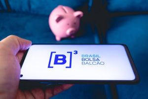 Celular com logo da B3, a bolsa brasileira, com cofre em formato de porquinho atrás