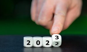 Em cima de uma mesa preta e com um fundo preto, dados com numerais formam o ano de 2022, e o último deles virando para o 3, ao ser manipulado por um homem.