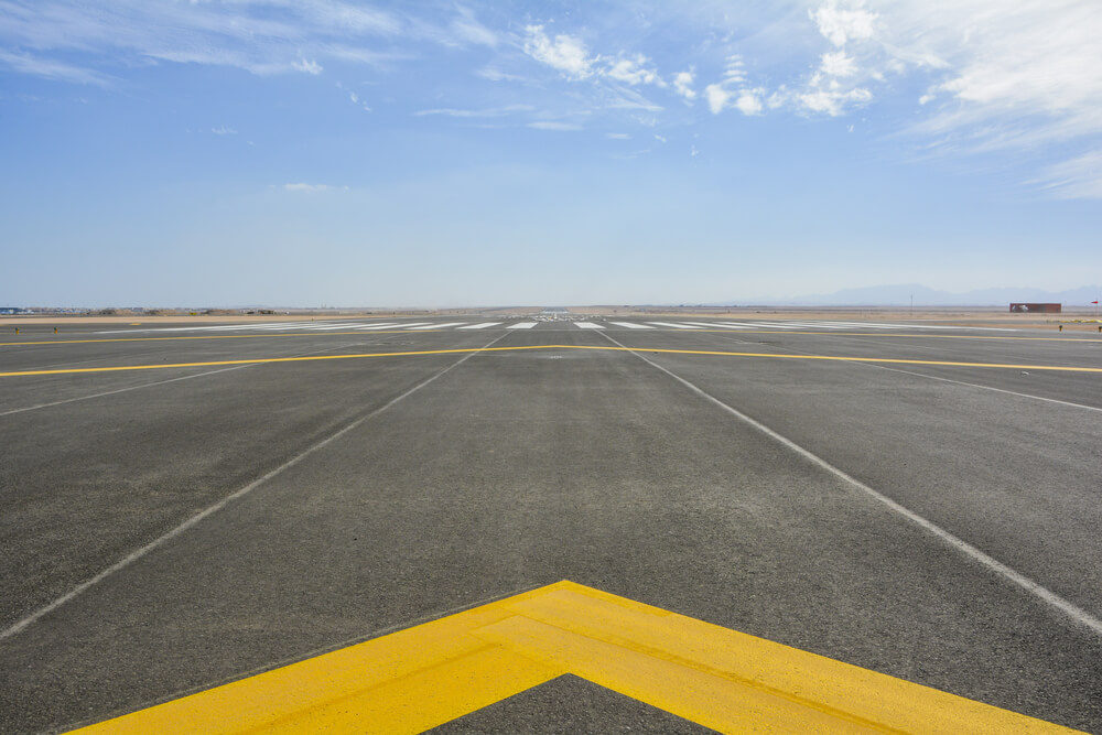 Imagem aproximada de uma pista de pouso de um aeroporto, sob a perspecitva do piloto, numa aterrisagem. Há também uma seta amarela no chão, indicando o sentido a ser percorrido.