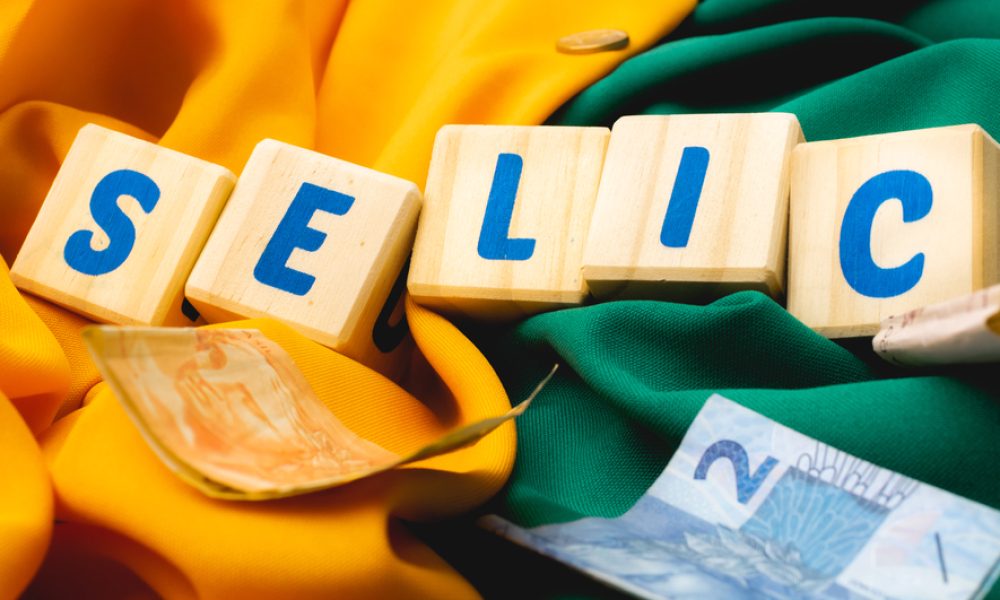 Blocos de letras formam a palavra Selic com notas de real ao redor e um fundo verde e amarelo