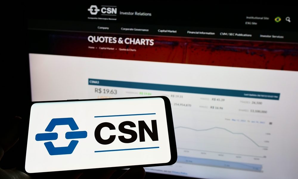 celular com logotipo da CSN na tela e, ao fundo, monitor com gráfico de cotação da ação da CSN